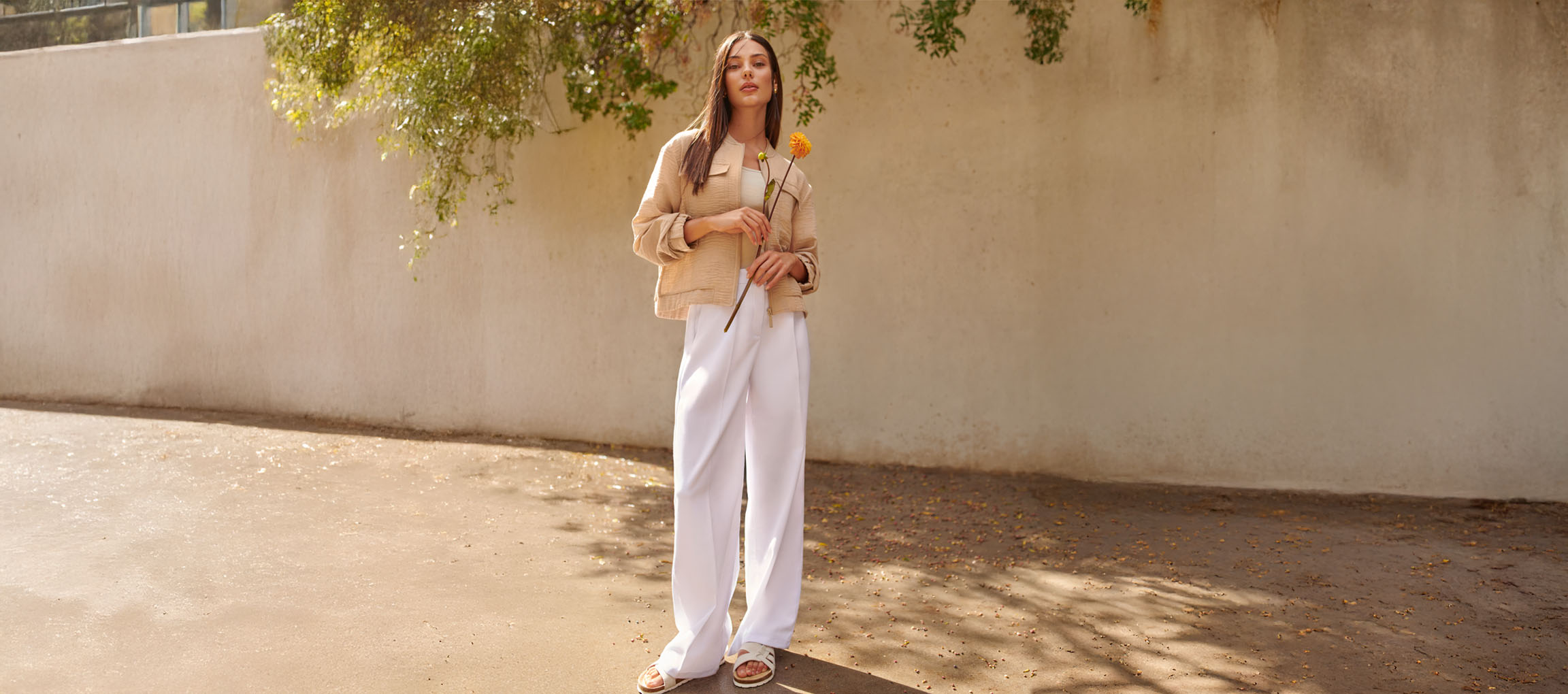 Sieviete smilškrāsas jakā, baltās biksēs un sandalēs, turot dzeltenu ziedu.