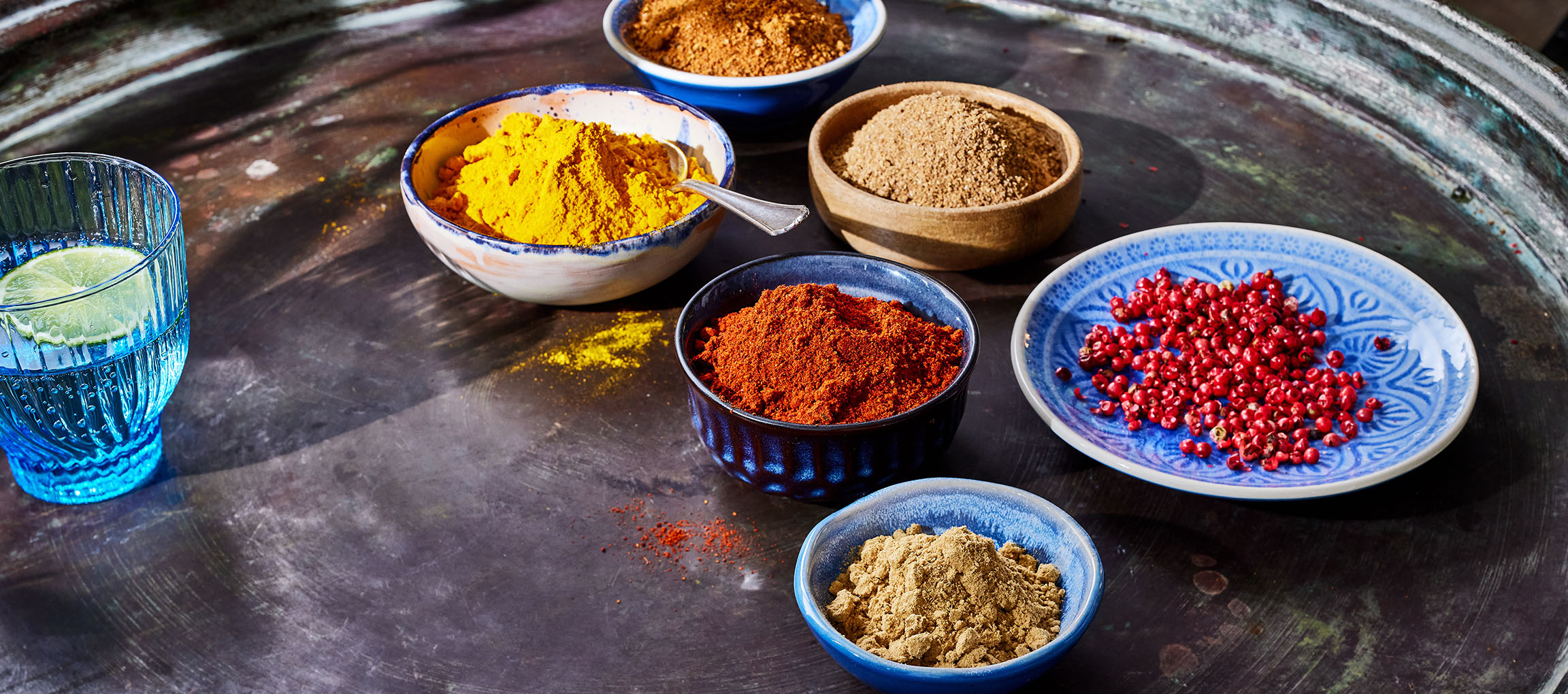 Dažādas garšvielas, piemēram, kurkuma, paprika un rozā pipari, bļodās uz tumšas virsmas.
