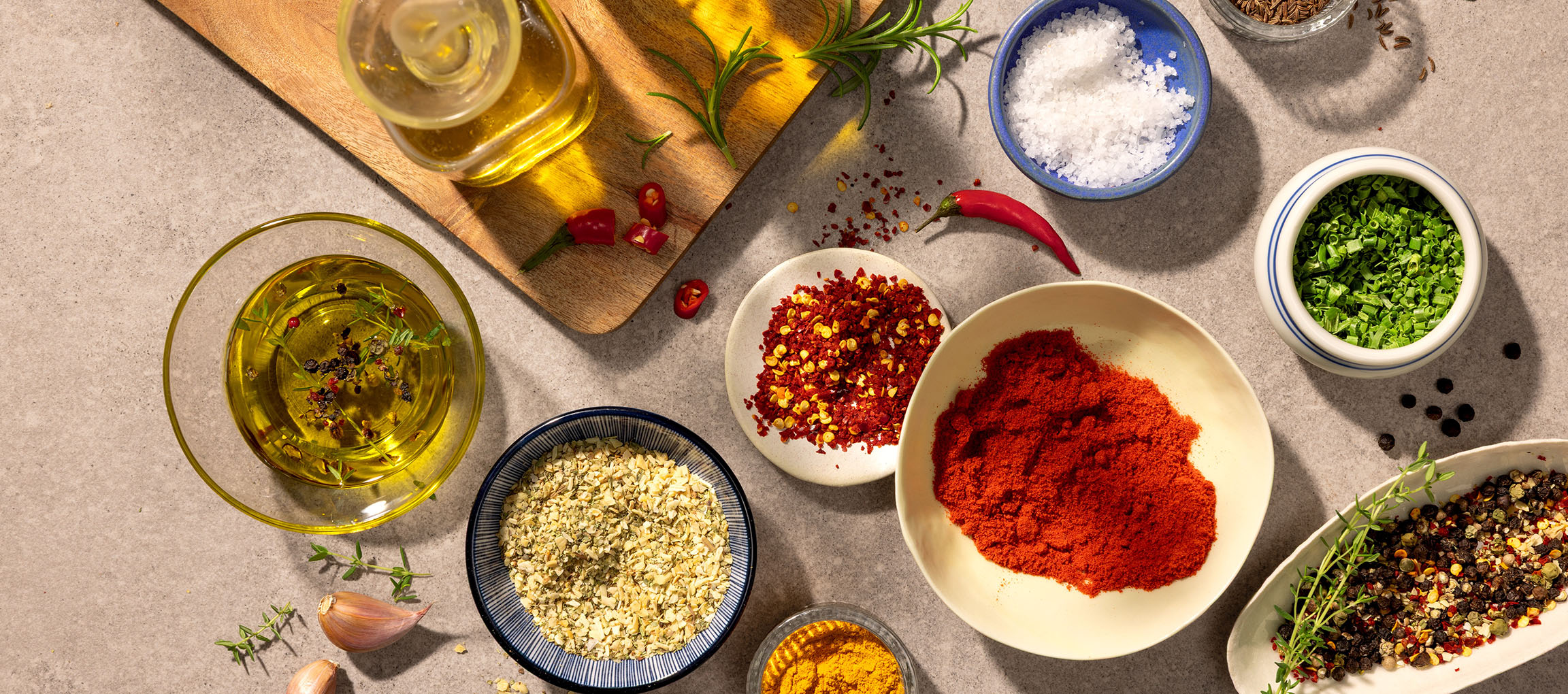 Dažādas garšvielas un eļļas, tostarp paprika, čili, sāls, pipari un olīveļļa, uz pelēka fona.