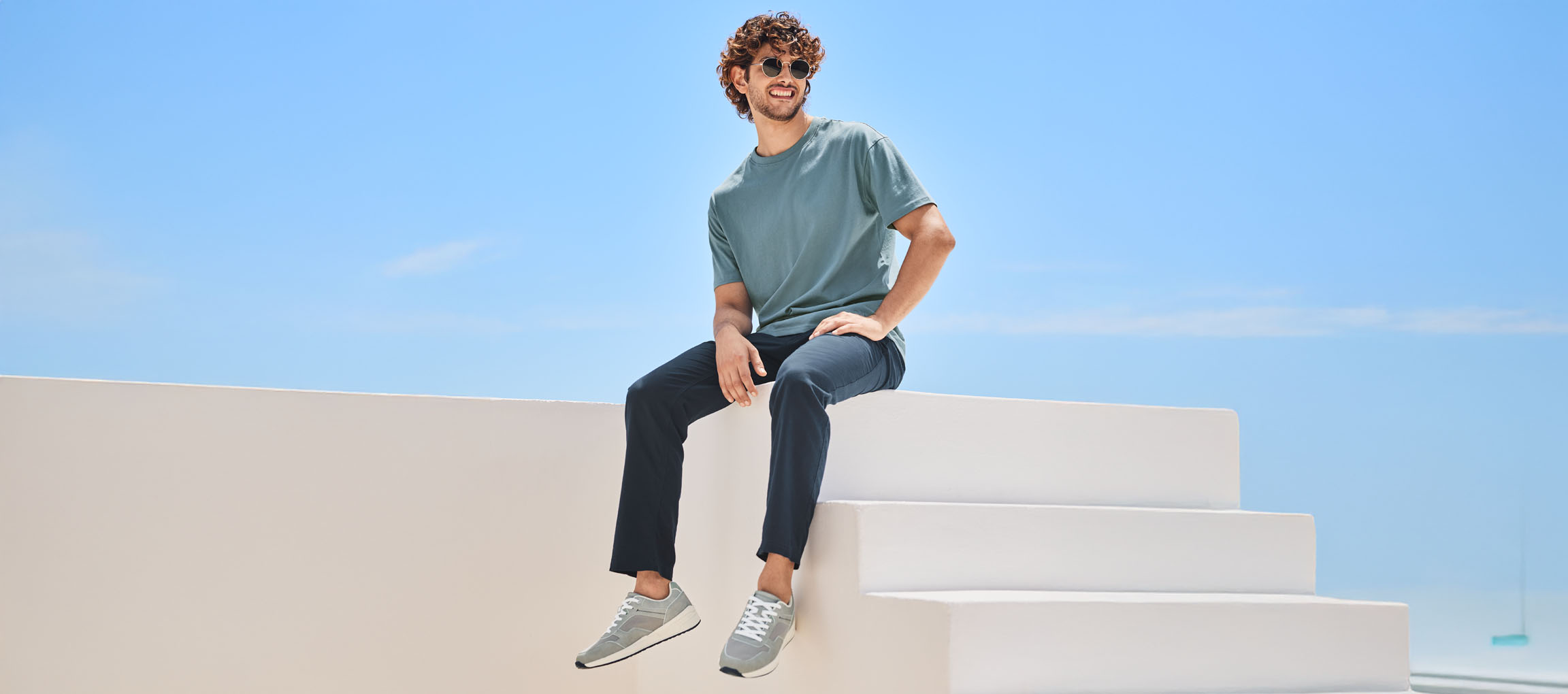 Vīrietis t-kreklā, biksēs un New Balance kedās, sēžot uz baltām kāpnēm.