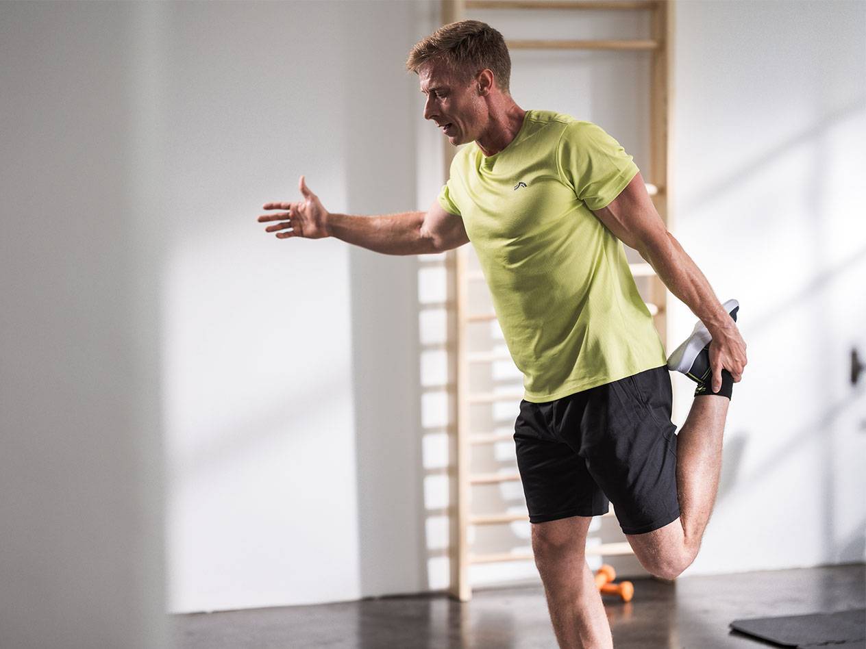 Мужчина растягивается в черных шортах и салатовой футболке в спортзале.