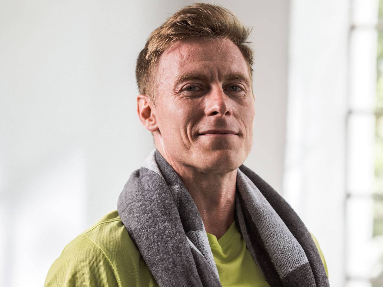 Светловолосый мужчина с полотенцем на шее смотрит в камеру.