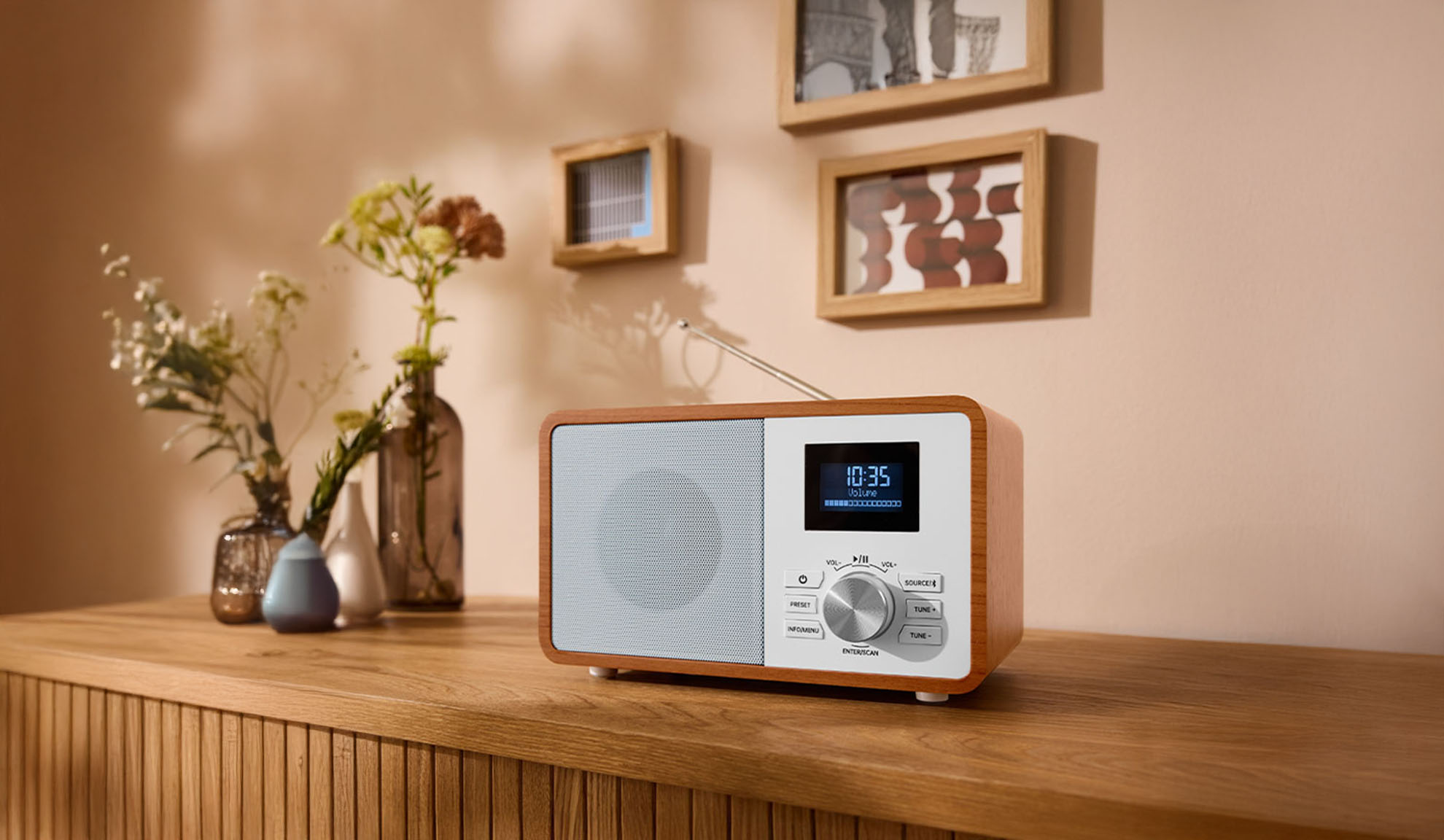 Koka digitālais radio uz kumodes, ar vāzēm un bilžu rāmjiem fonā.