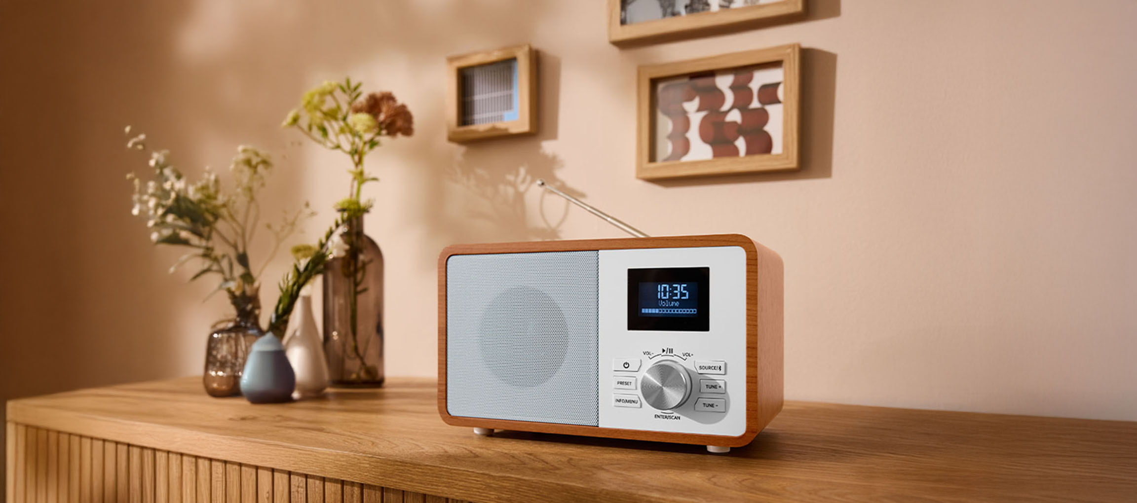 Koka digitālais radio uz kumodes, ar vāzēm un gleznām pie sienas.
