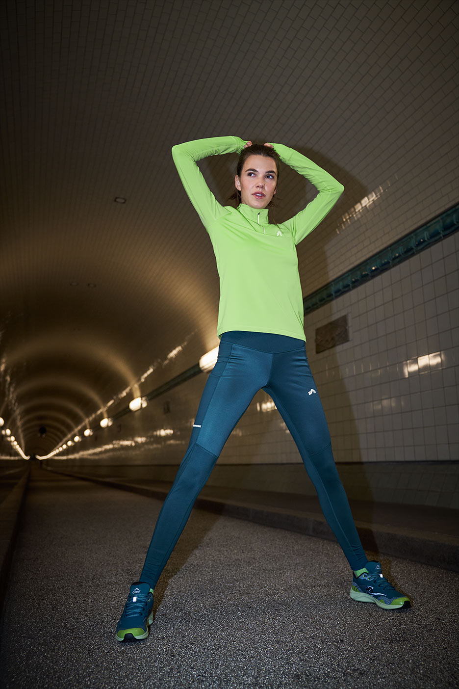 Sieviete laima krāsas sporta jakā un tumši zilās legingos iesildās tunelī.