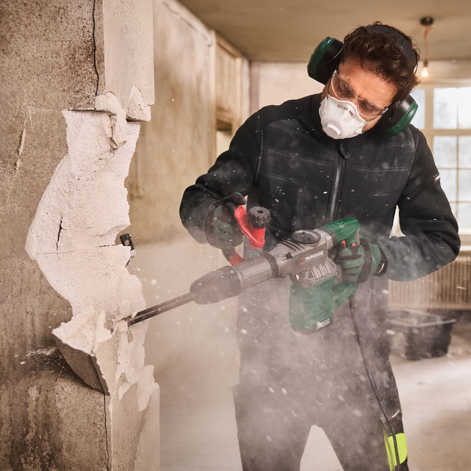 Vīrietis ar laužamām āmuru, masku un ausu aizsargiem nojauc sienu.