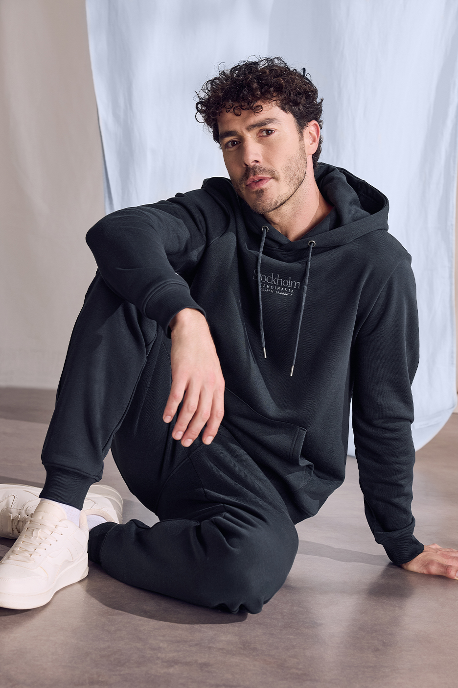 Vīrietis tumši zilā džemperī ar kapuci ar uzrakstu 'Stockholm Scandinavia' un sporta biksēs.