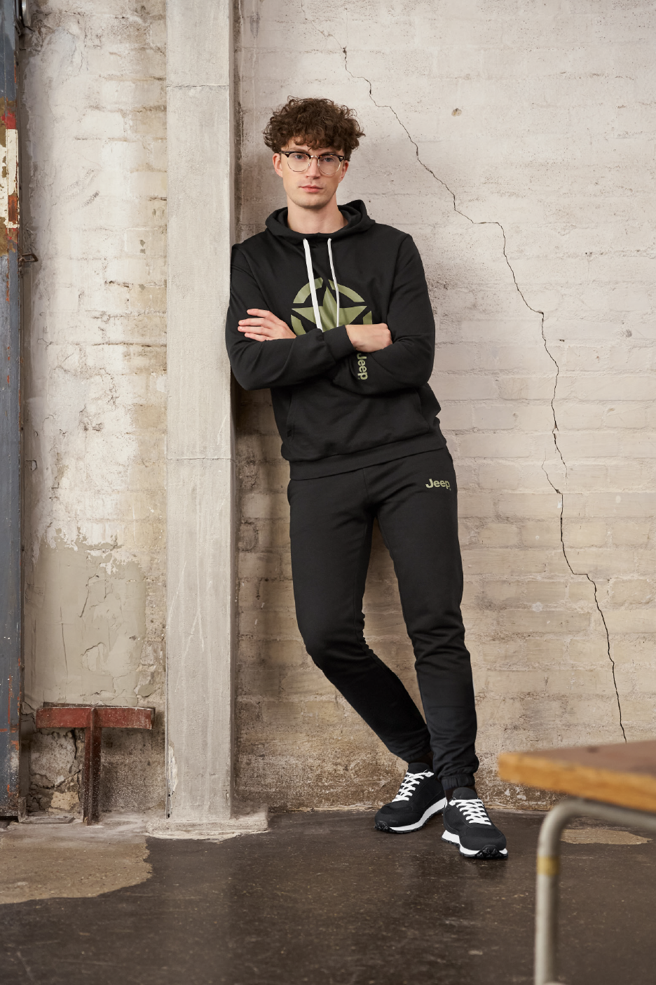 Vīrietis melnā sporta tērpā ar kapuci un biksēm, kas atspiedies pret sienu.
