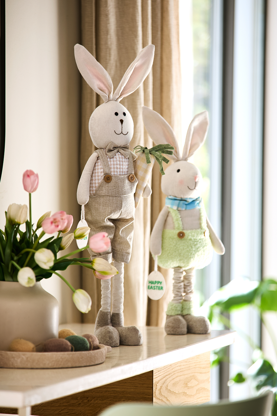 Divas Lieldienu zaķu rotaļlietas un vāze ar tulpēm uz galda.