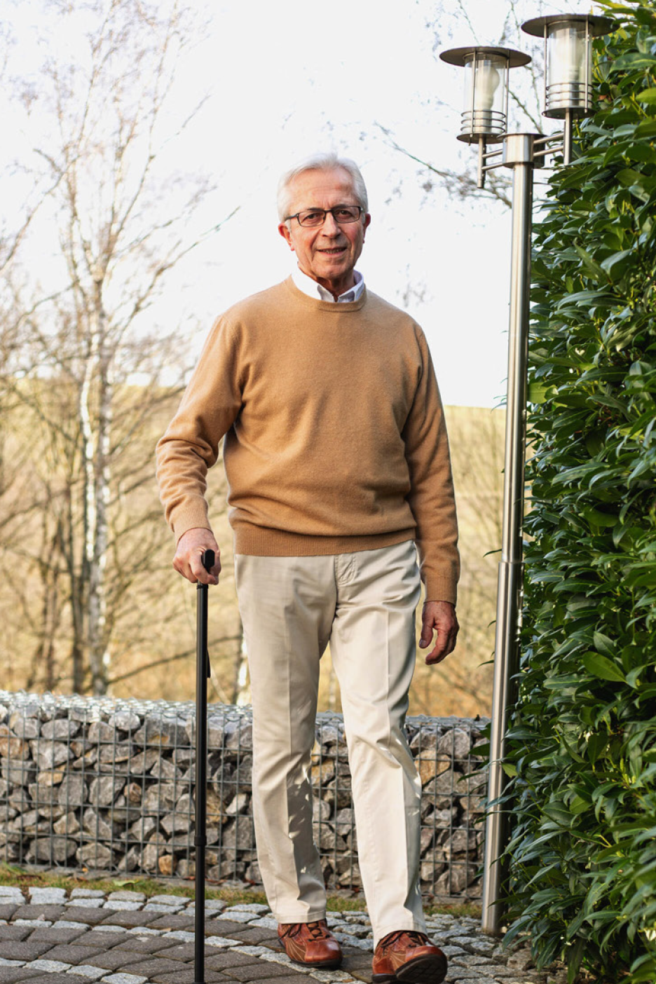 Vecāks vīrietis bēšā džemperī un gaišās biksēs, atbalstoties uz spieķa, smaida.