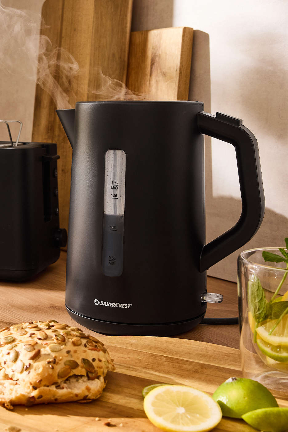 Melna Silvercrest tējkanna ar tvaiku, tosteris, maizīte un citrons uz koka dēlīša.