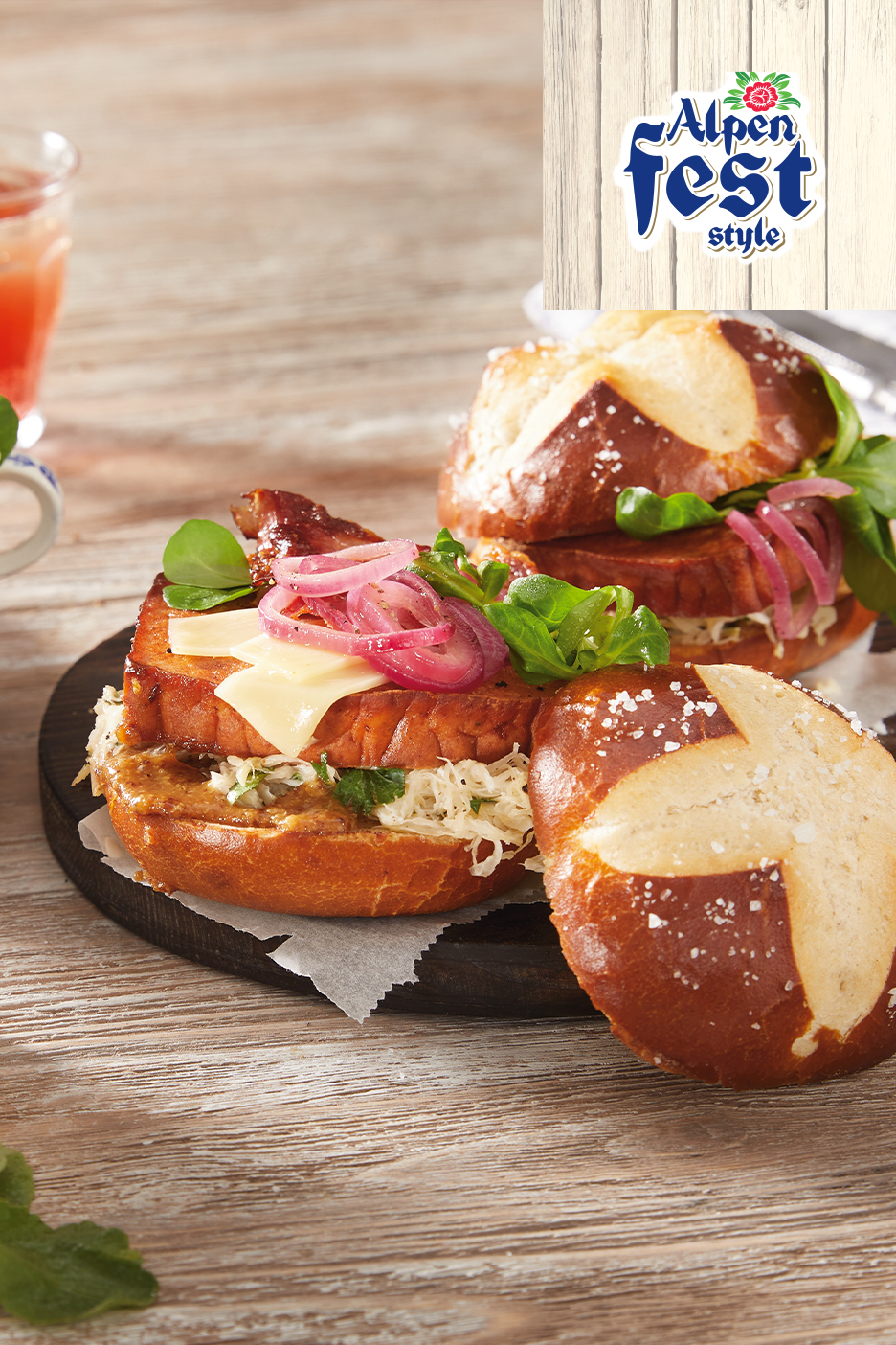 Divas maizītes ar gaļu, sieru, marinētiem sīpoliem un kāpostiem uz koka galda. Augšējā labajā stūrī ir uzlīme ar uzrakstu 'Alpen Fest style'.