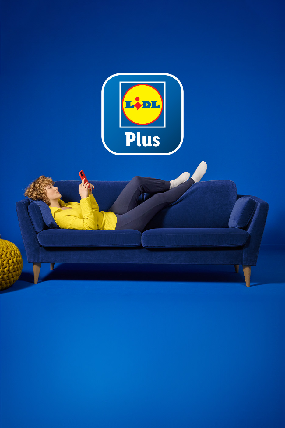 Sieviete uz zila dīvāna skatās telefonā, virs viņas Lidl Plus logotips.