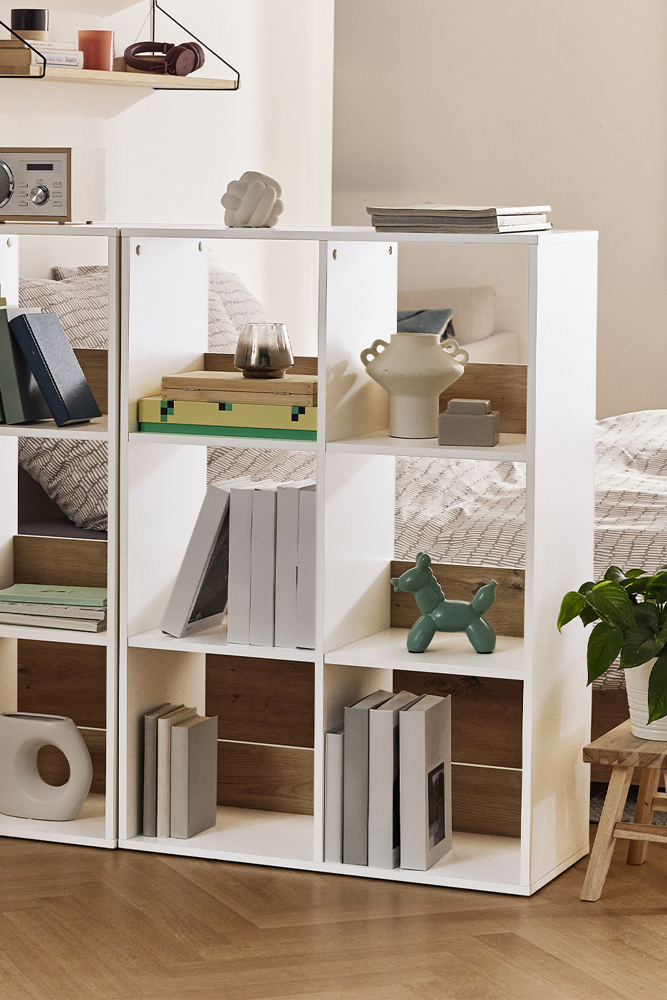 Balts plaukts ar grāmatām, vāzēm un dekoratīviem priekšmetiem, kas atdala guļamistabas zonu.