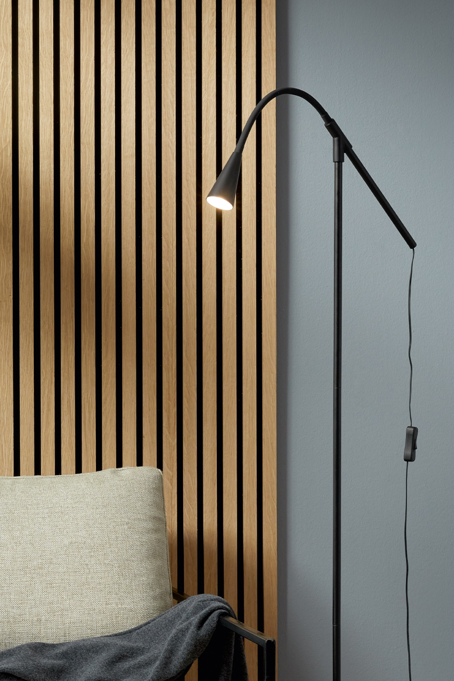Melna stāvlampa ar izliektu augšdaļu un gaišs spilvens uz krēsla pie koka paneļu sienas.