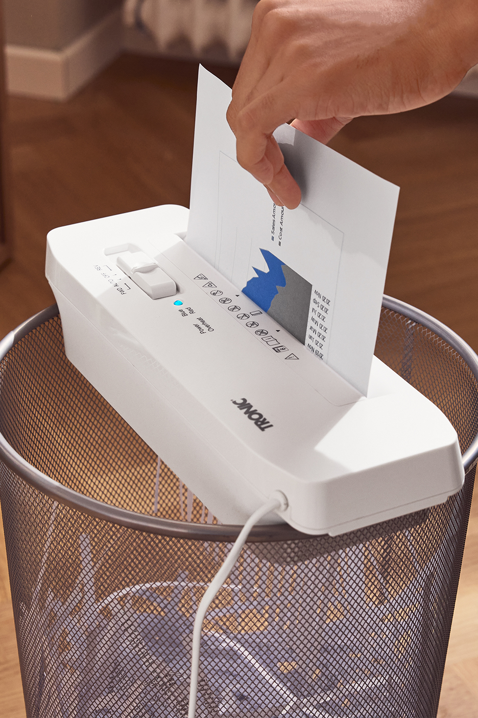 Roka ievieto dokumentu ar diagrammu baltā TRONIC papīra smalcinātājā, kas novietots uz atkritumu tvertnes.