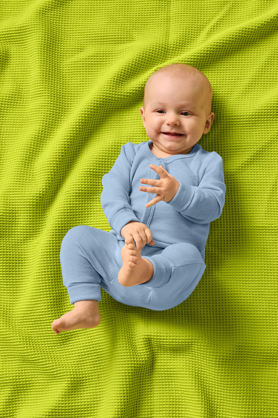 Zīdainis gaiši zilā kombinezonā guļ uz spilgti zaļas vafeļu segas.