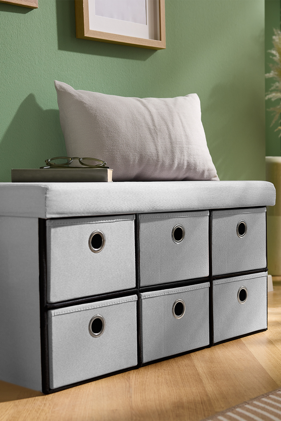 Pelēks uzglabāšanas sols ar sešām atvilktnēm un spilvenu telpā ar zaļu sienu.