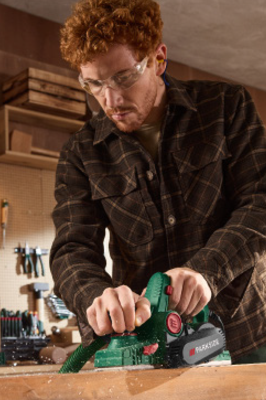 Vīrietis aizsargbrillēs un ausu aizbāžņos ēvelē koka dēli ar ēveli.