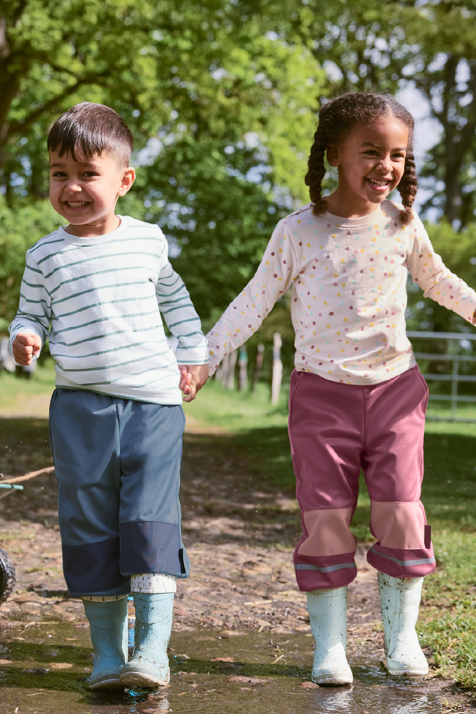 Divi bērni ūdensnecaurlaidīgās biksēs un gumijas zābakos turas pie rokām un smaida, stāvot peļķē.