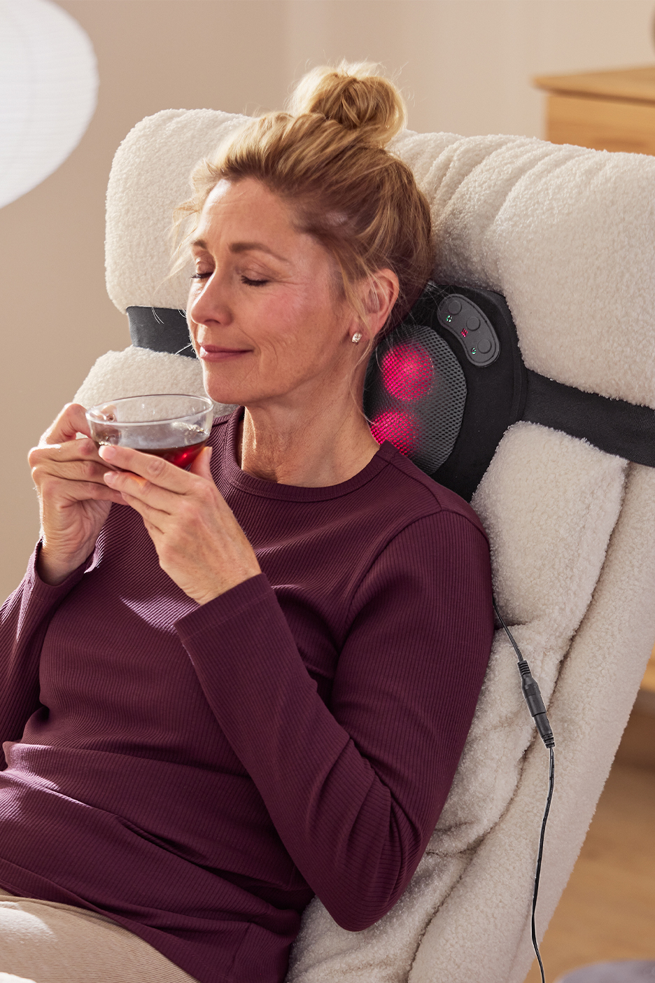 Женщина наслаждается чаем, сидя в кресле с массажной подушкой на шее.