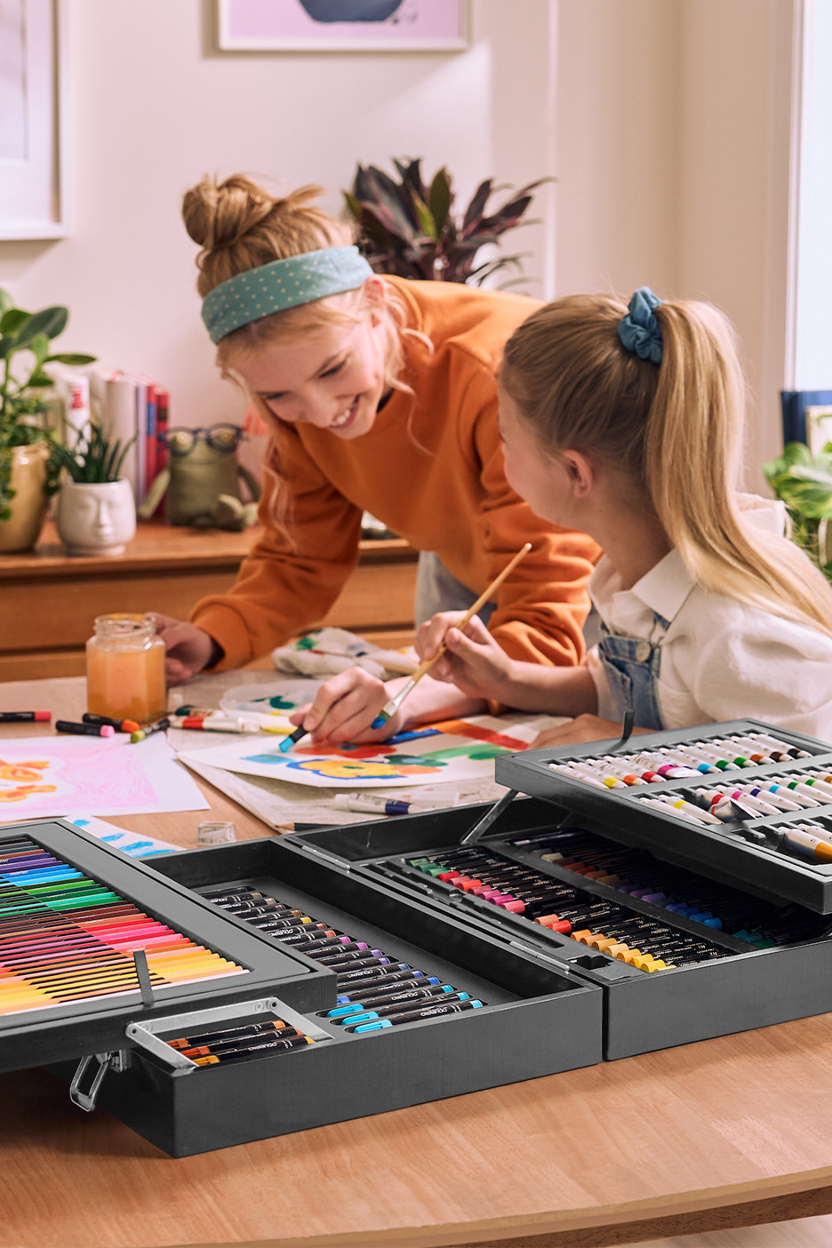 Две девочки рисуют за столом, используя большой набор для творчества с карандашами и красками.