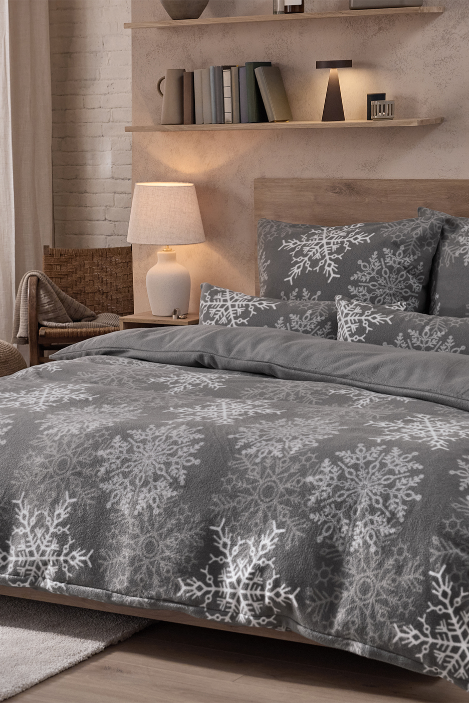 Pelēka gultasveļa ar baltām sniegpārslām guļamistabā ar koka plauktiem un lampām.