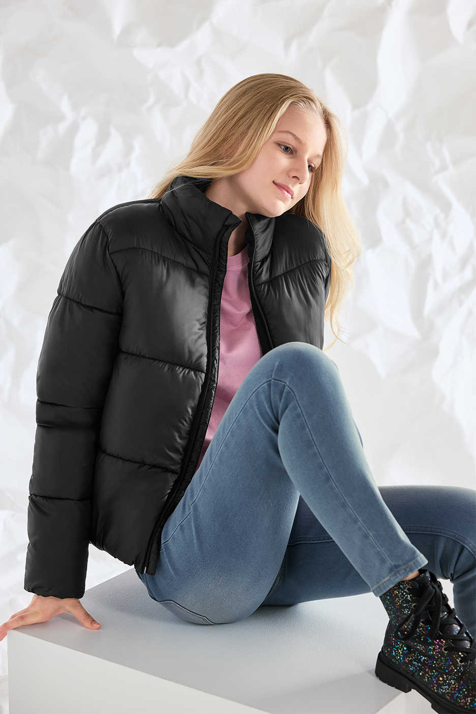 Meitene melnā pufīgā jakā, rozā T-kreklā, džinsos un spīdīgos zābakos.