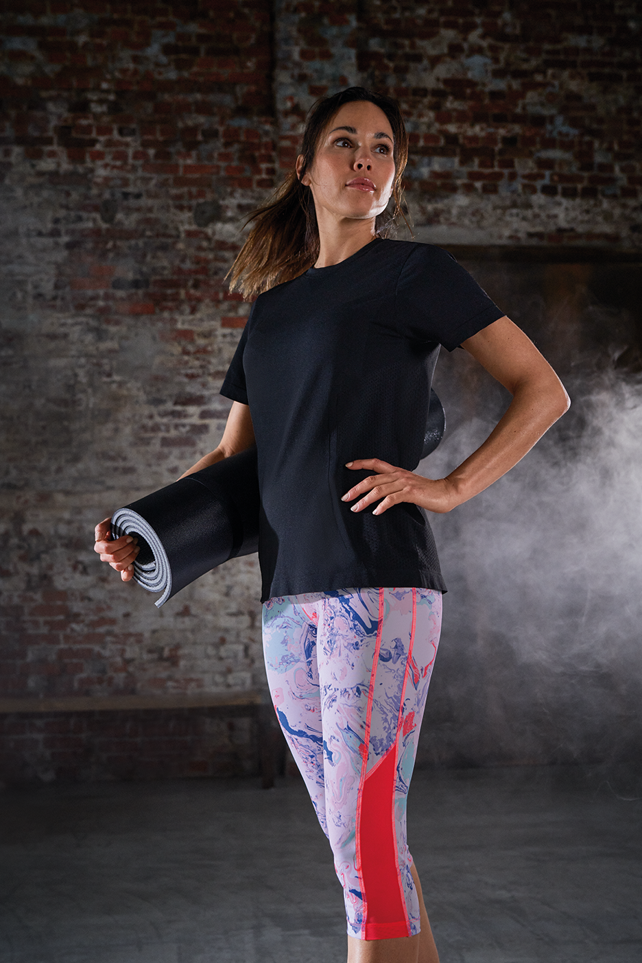 Sieviete melnā T-kreklā un rakstainās legingos tur jogas paklājiņu.