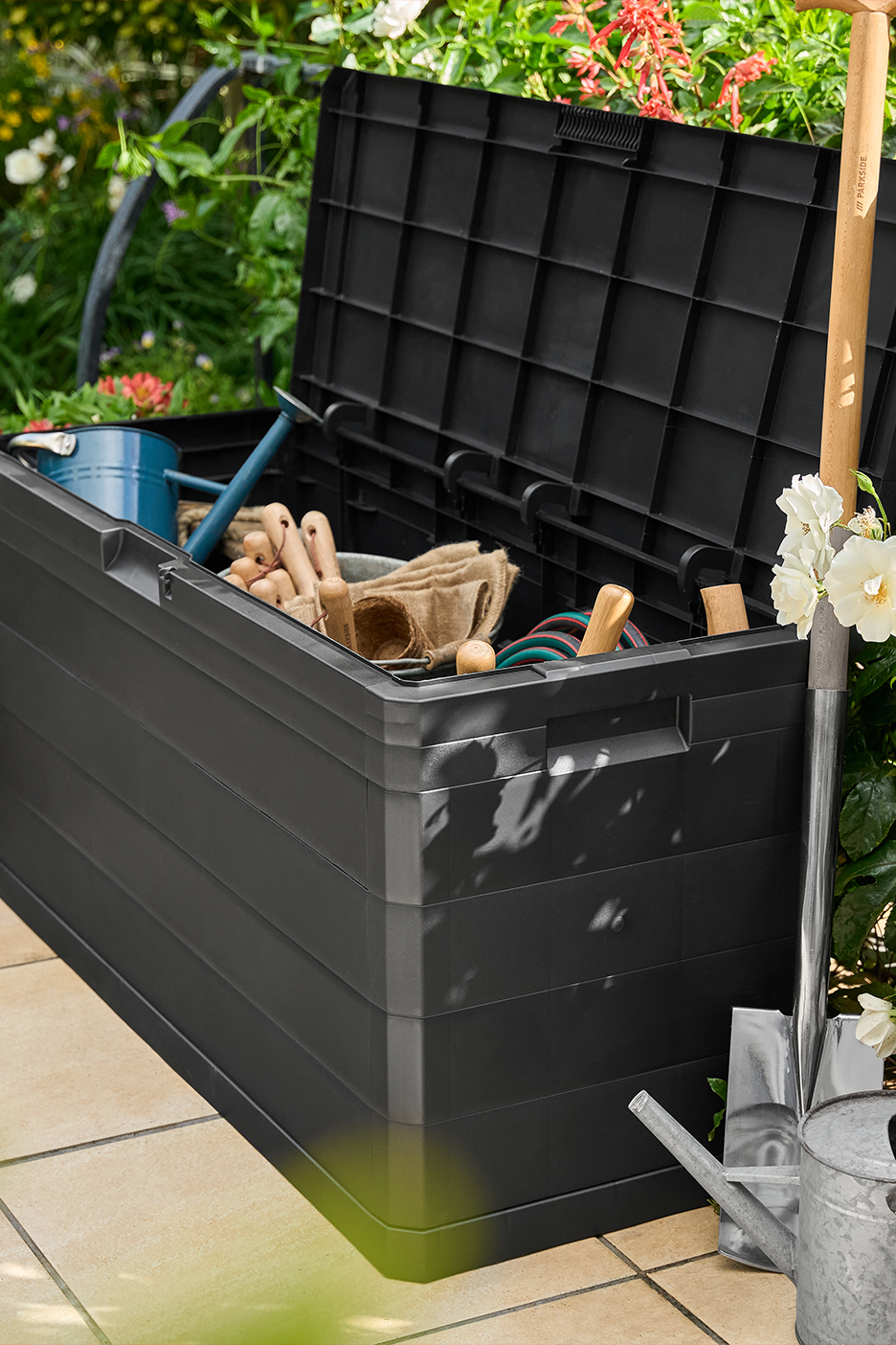 Melna dārza uzglabāšanas kaste ar atvērtu vāku, piepildīta ar dārza instrumentiem, laistīšanas kannu un lāpstu.