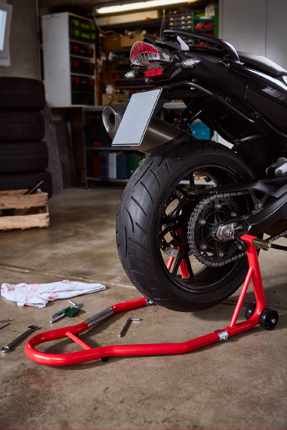 Motocikls uz sarkana statīva garāžā ar instrumentiem un riepām.