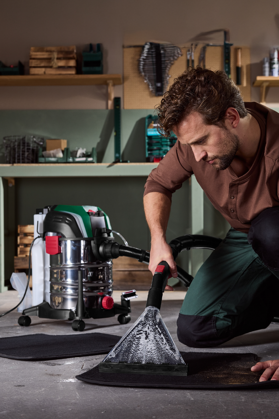 Vīrietis tīra automašīnas paklājiņu ar mitrās tīrīšanas putekļsūcēju darbnīcā.