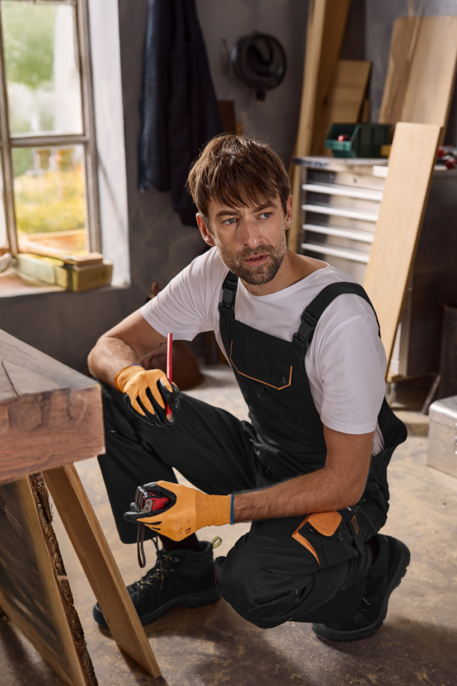 Vīrietis darba apģērbā un cimdos mēra koka dēli.