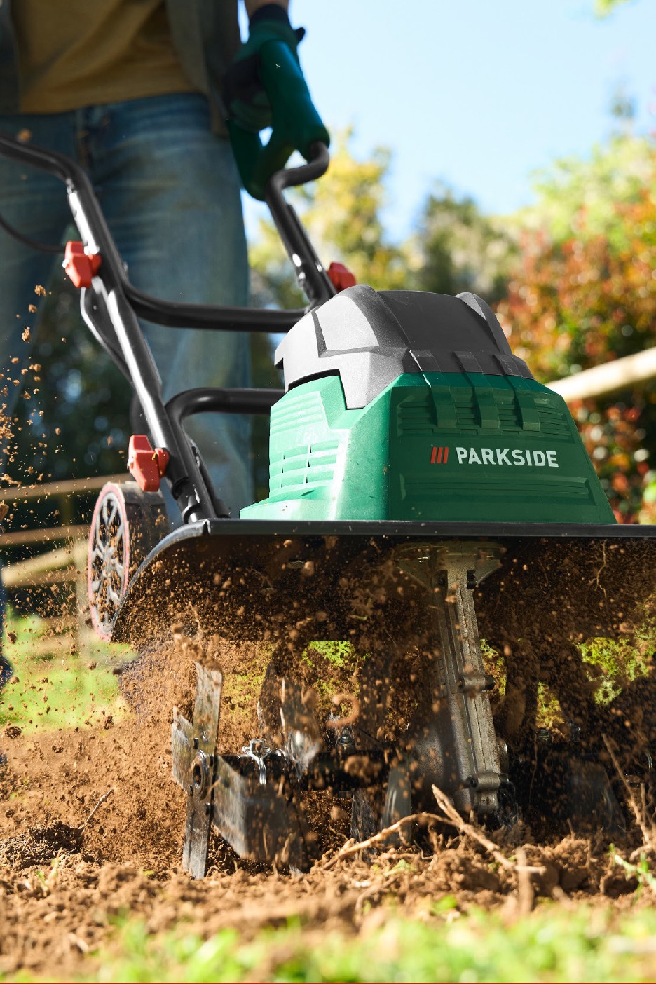 Cilvēks džinsos un cimdos izmanto zaļu kultivatoru, kas apstrādā zemi.