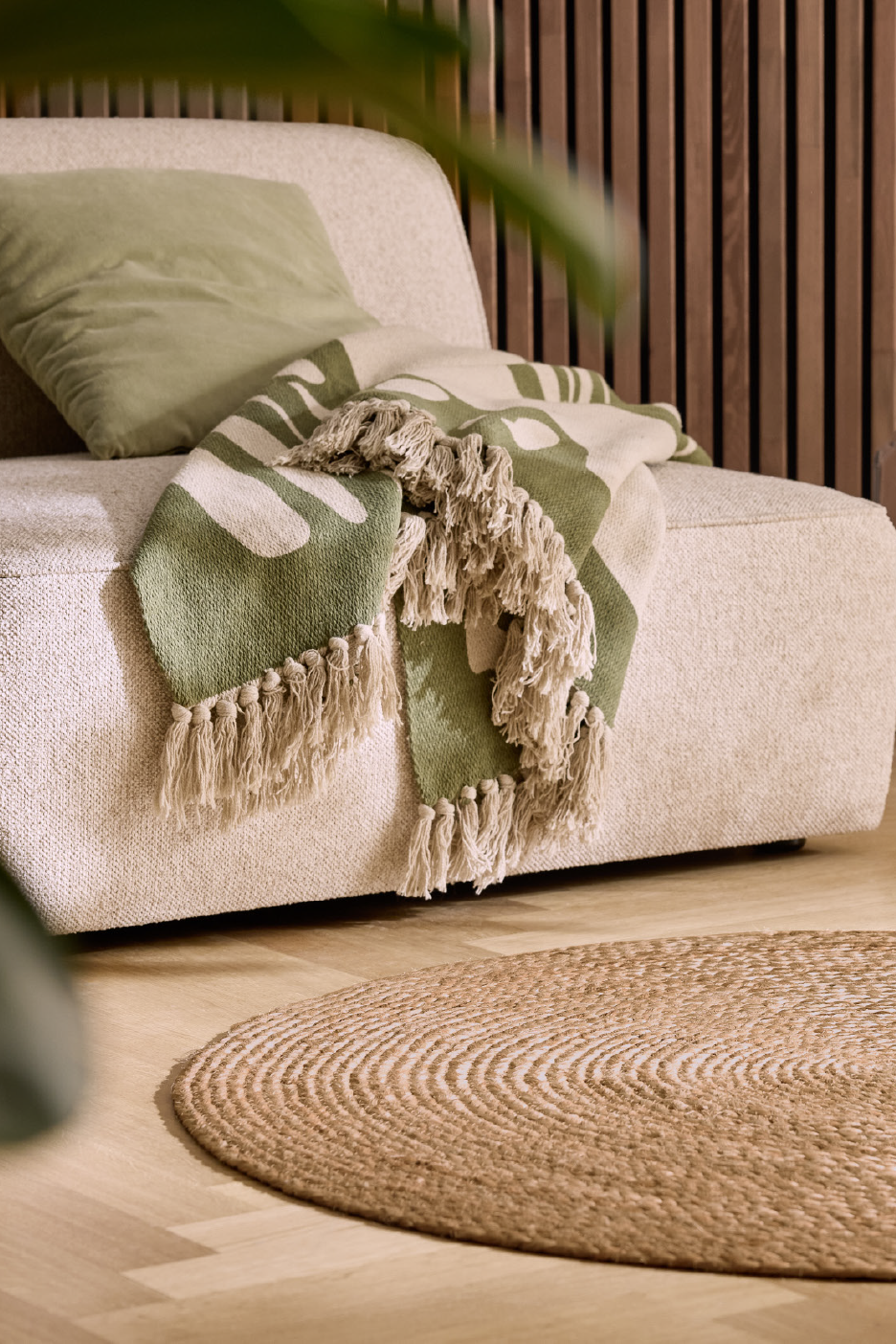 Bēšs dīvāns ar zaļu spilvenu un pledu, uz grīdas apaļš džutas paklājs.