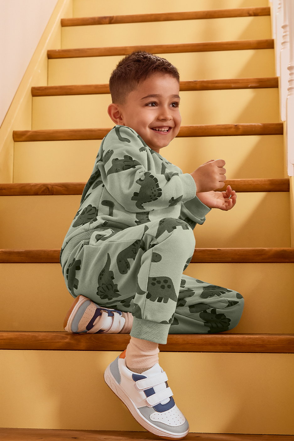 Zēns zaļā sporta tērpā ar dinozauriem sēž uz kāpnēm.