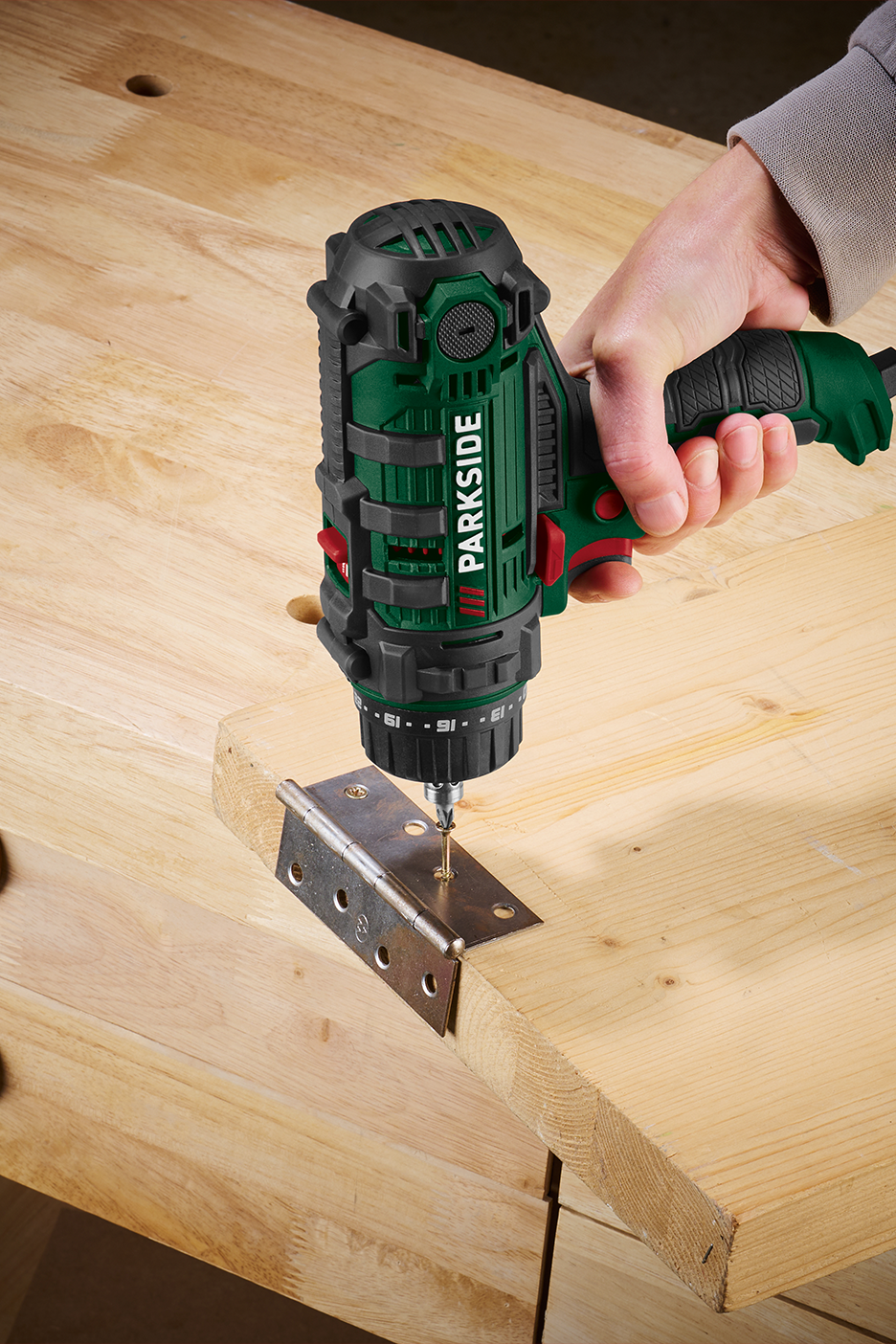 Cilvēks izmanto urbjmašīnu-skrūvgriezi, lai piestiprinātu eņģi pie koka dēļa.