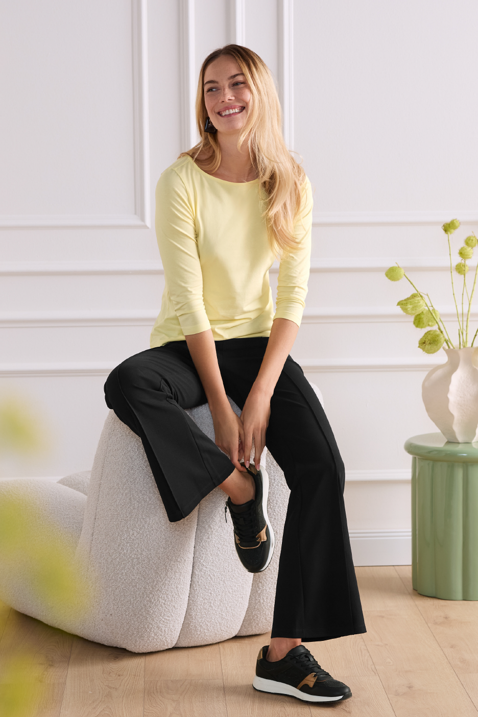 Sieviete dzeltenā blūzē un melnās biksēs sēž uz pufa, smaida un skatās uz sāniem.