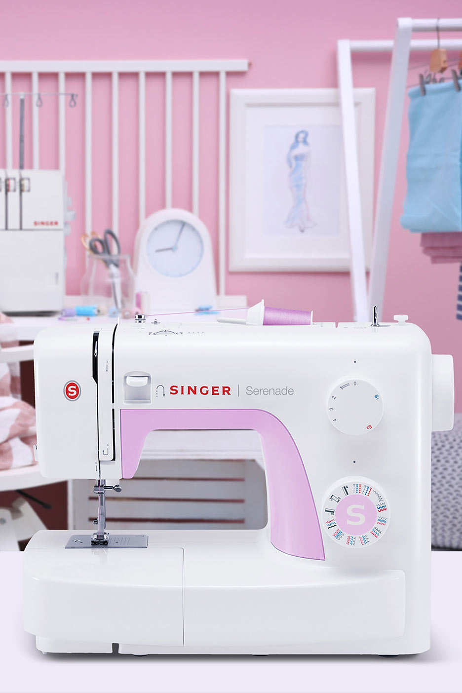 Balta šujmašīna ar rozā akcentiem uz galda rozā telpā ar šūšanas piederumiem.
