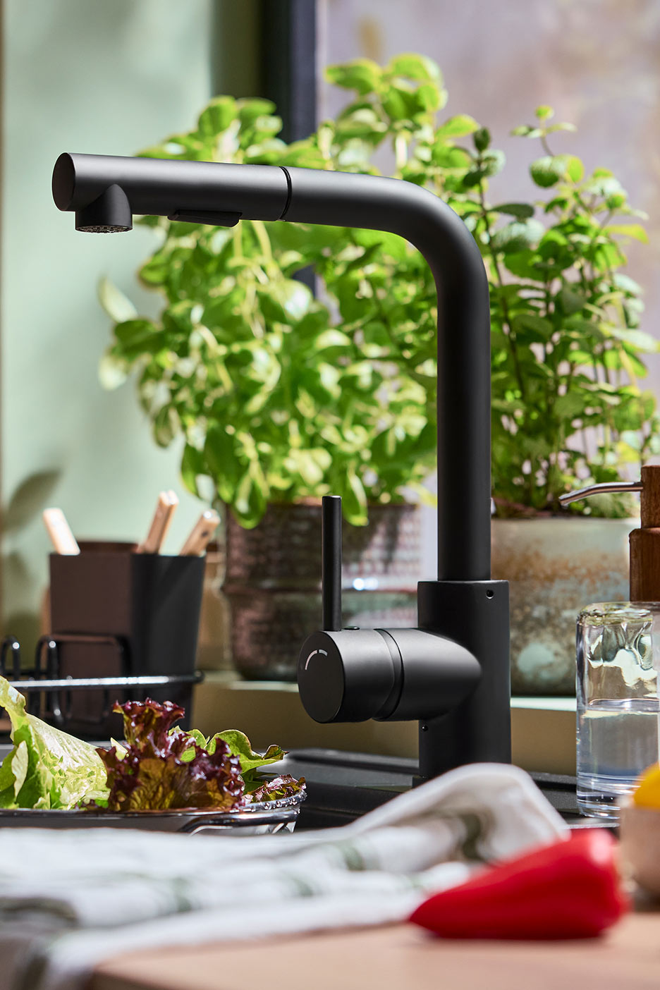 Melns virtuves jaucējkrāns ar izvelkamu dušu, salāti un dārzeņi uz galda.