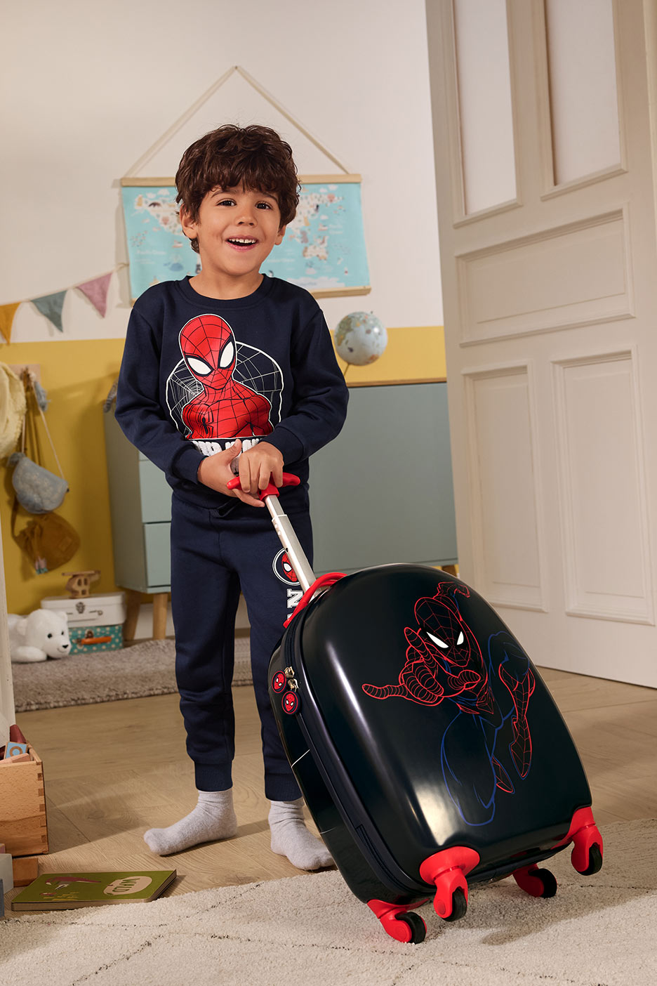 Zēns ar zirnekļcilvēka tērpu un koferi, kas stāv uz paklāja.