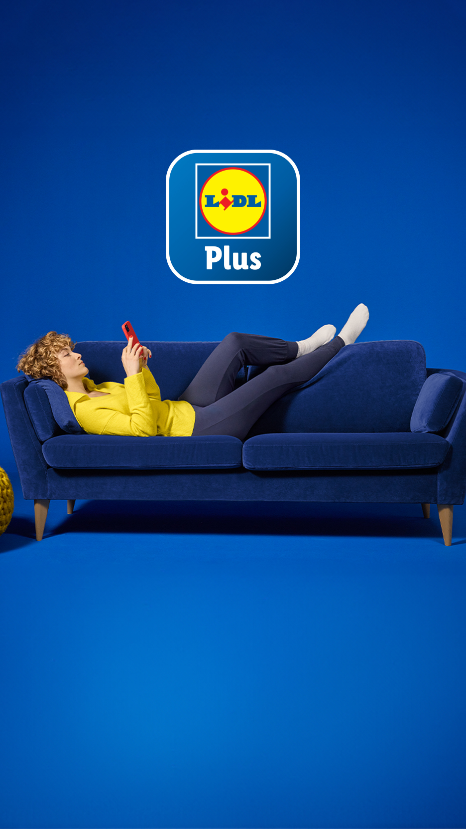 Sieviete atpūšas uz dīvāna, lietojot tālruni, ar Lidl Plus logotipu fonā.