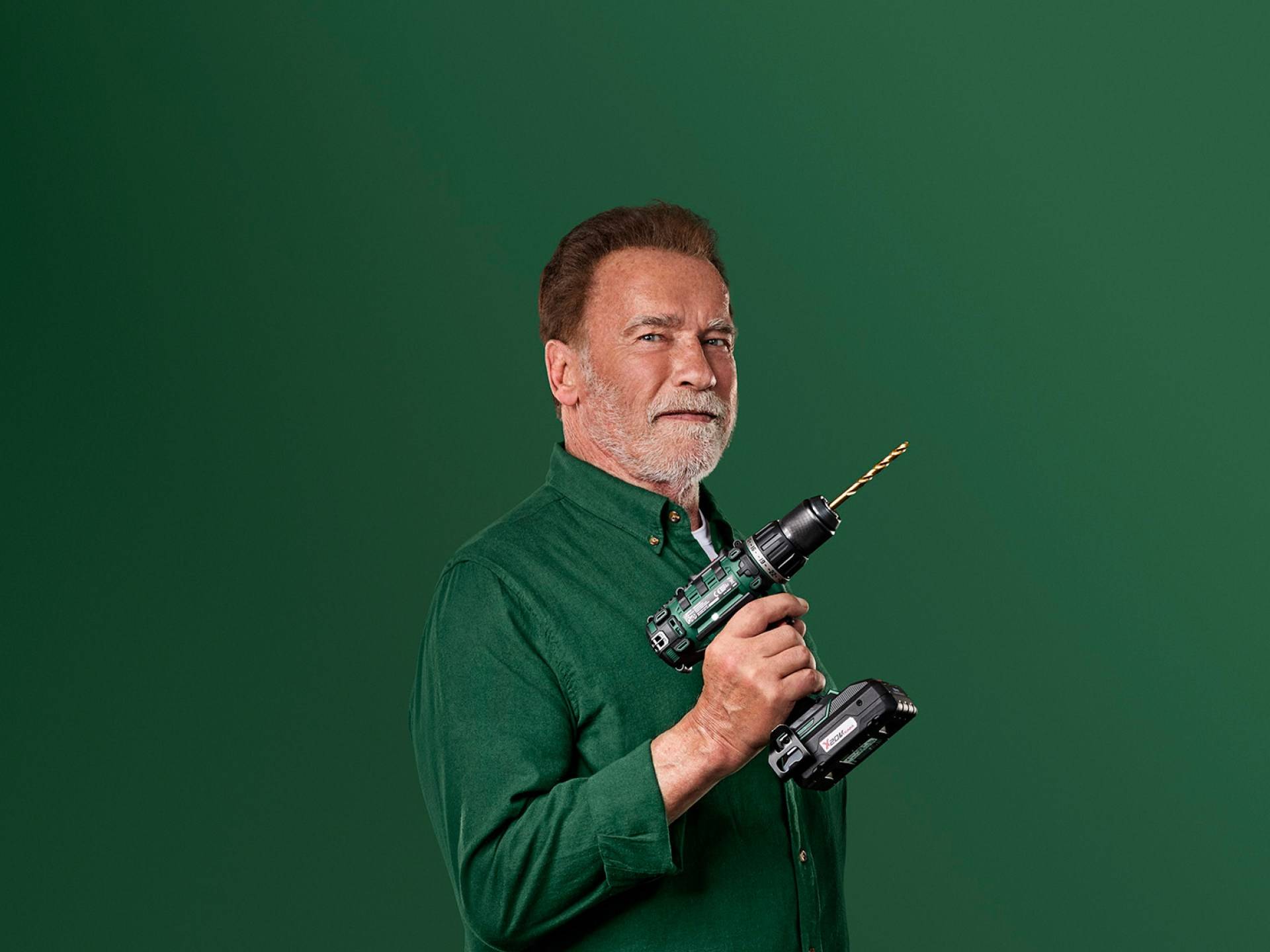Arnolds Švarcenegers tur zaļu elektrisko urbi uz zaļa fona.