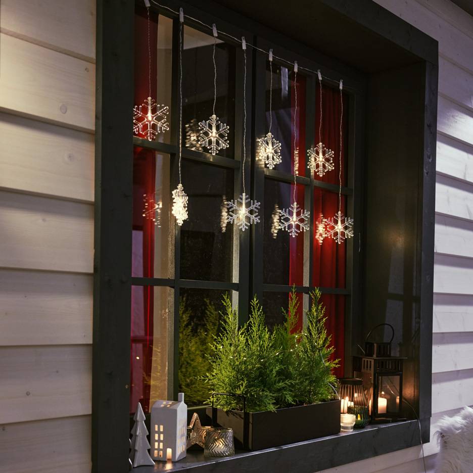 Гирлянда из снежинок в окне с маленькими елями и свечами на подоконнике.