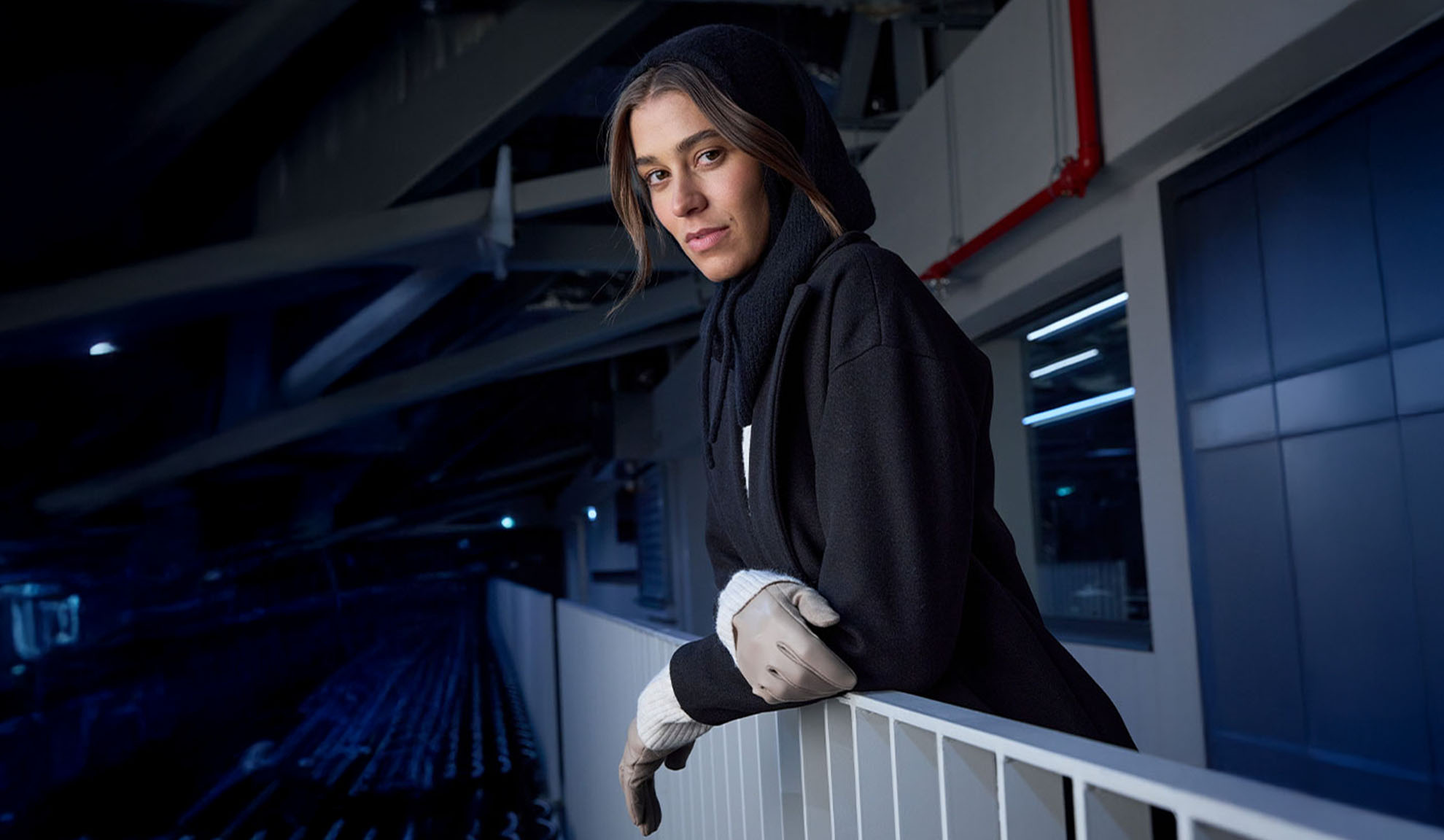 Женщина в черном пальто и капюшоне, опирающаяся на перила в современном интерьере.