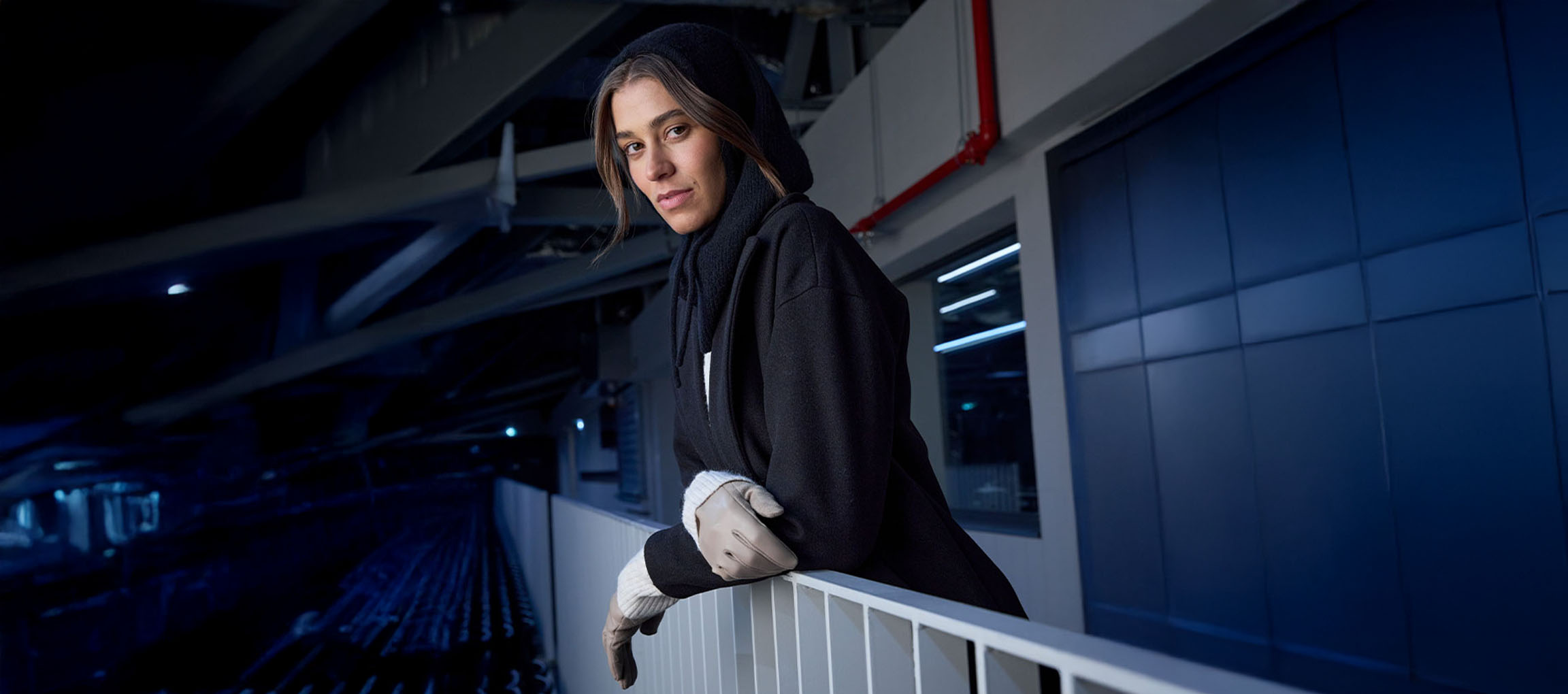 Женщина в черном пальто и перчатках опирается на перила в городской среде.