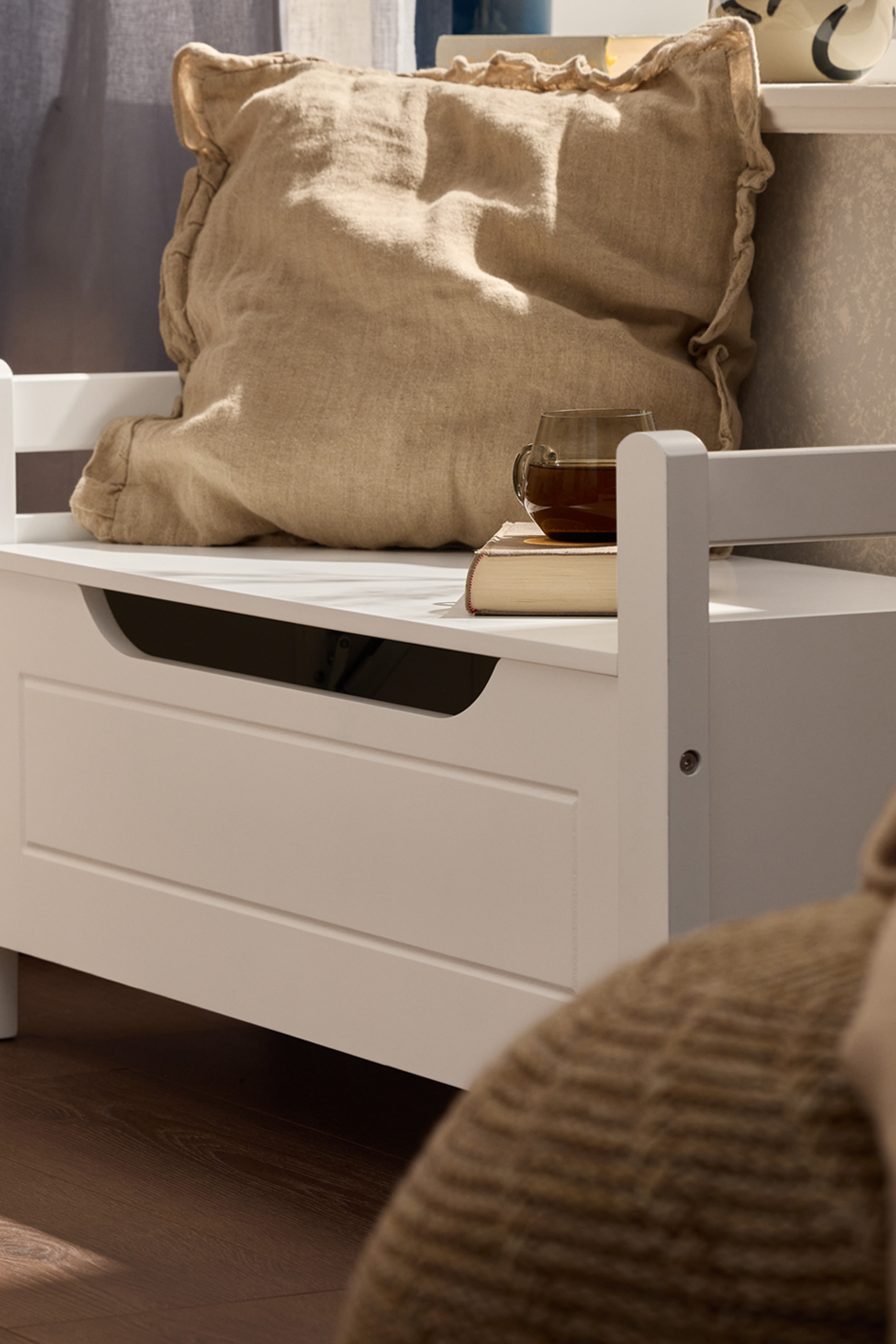 Белая деревянная скамья с подушкой, книгой и чашкой чая на темном деревянном полу.