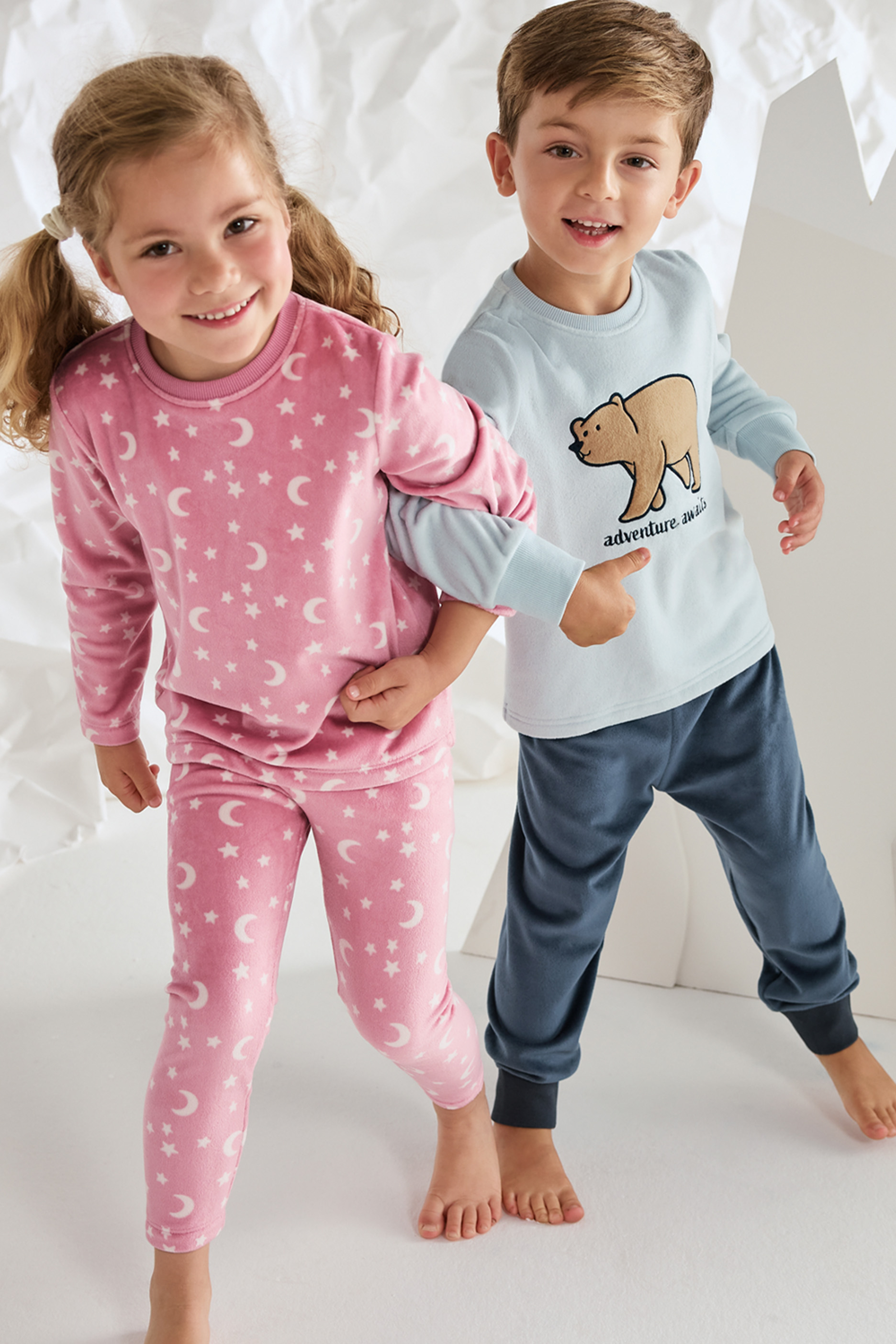 Двое детей в пижамах: девочка в розовой с принтом луны и звезд, мальчик в голубой с медведем.
