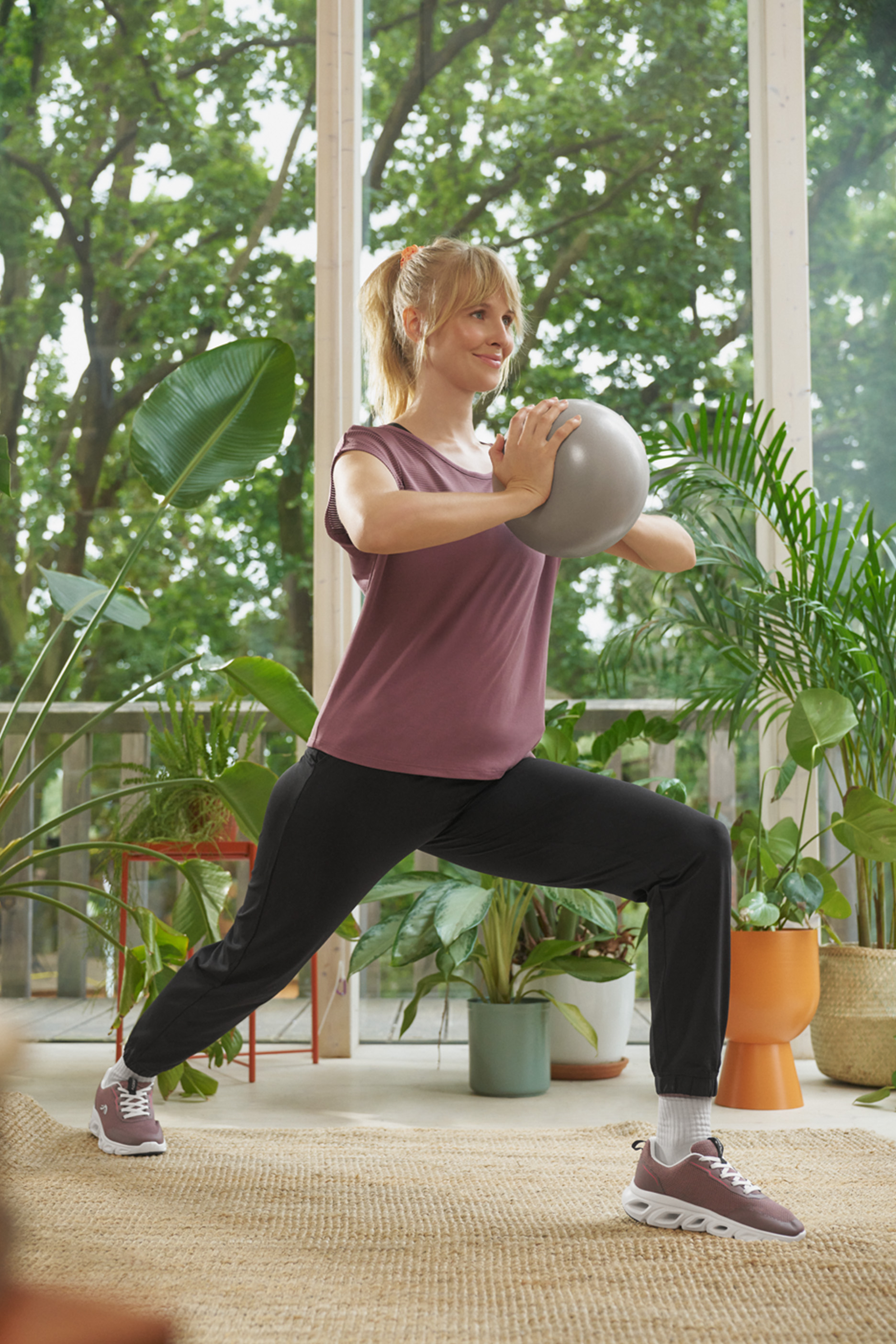 Женщина тренируется с маленьким мячом в спортивной одежде, окруженная зелеными растениями.