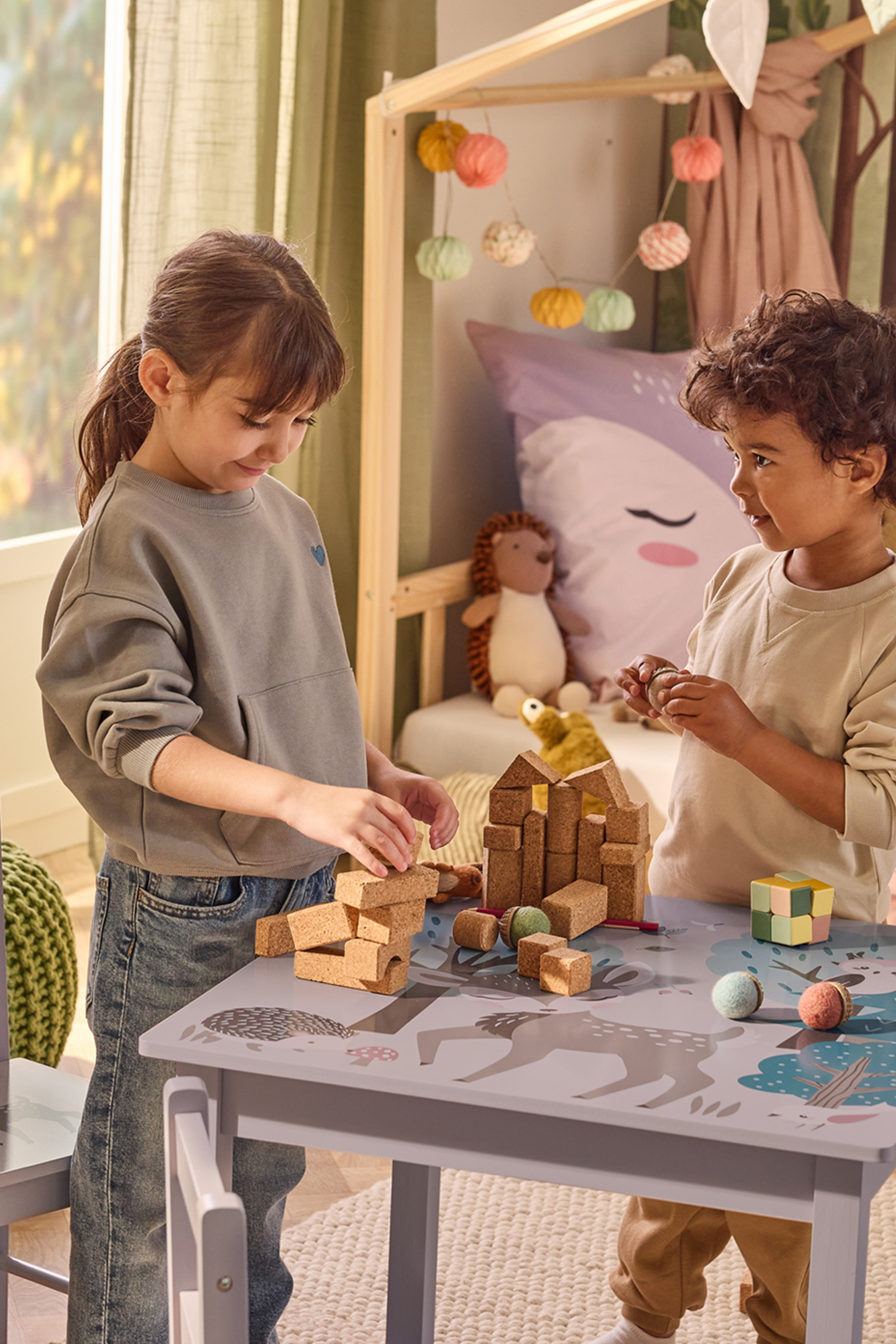 Divi bērni spēlējas ar korķa klučiem un rotaļlietām uz galda ar meža dzīvnieku apdruku.